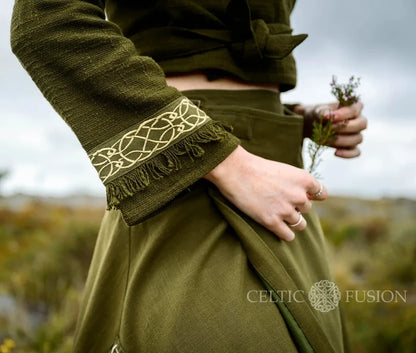 Close-up of the olive green folklore wrap top showing cream Celtic knotwork embroidery and soft fringe on the flared cuff.