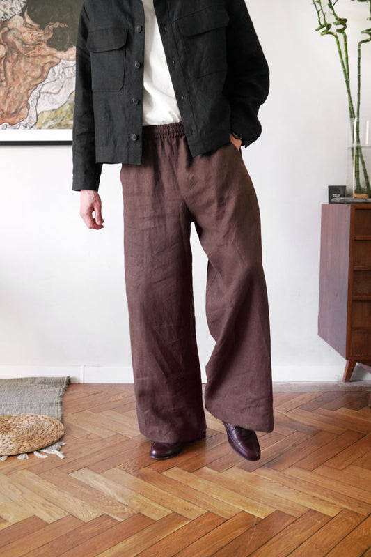 Person wearing a dark jacket and brown wide-leg pants standing in a room with wooden flooring.