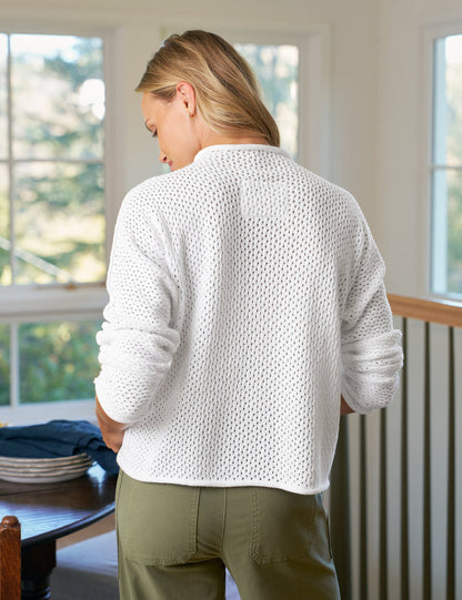 Person wearing a white textured sweater and green pants standing in a room with large windows.
