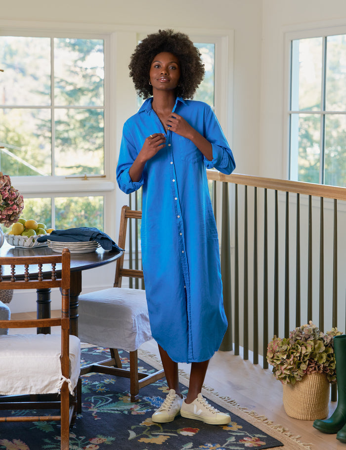 Woman wearing a blue dress standing in a room with large windows and furniture.